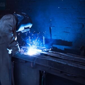 Industrial worker welding metal indoors, emitting bright blue sparks and smoke.